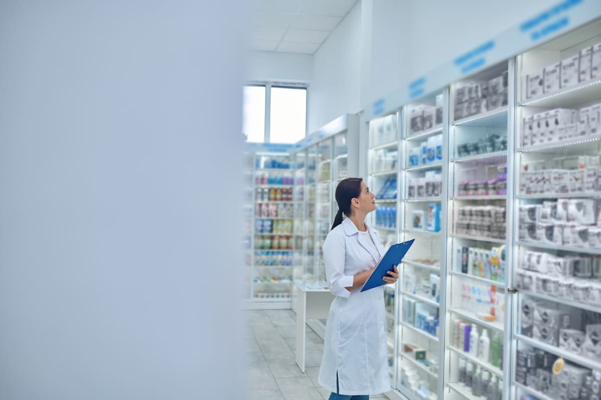 Farmacêutica analisando prateleiras em uma farmácia, verificando o armazenamento de medicamentos para controle e organização dos produtos.