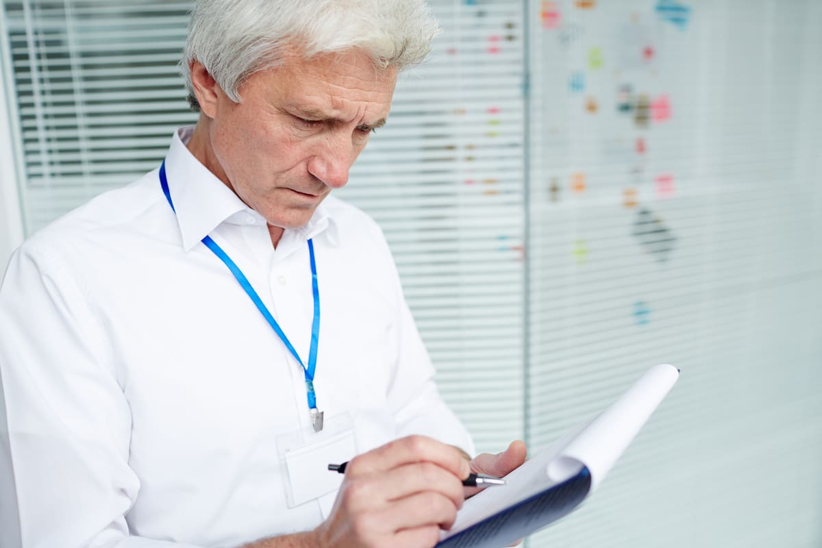 Profissional com crachá e camisa branca anota dados em uma prancheta, realizando uma auditoria hospitalar.